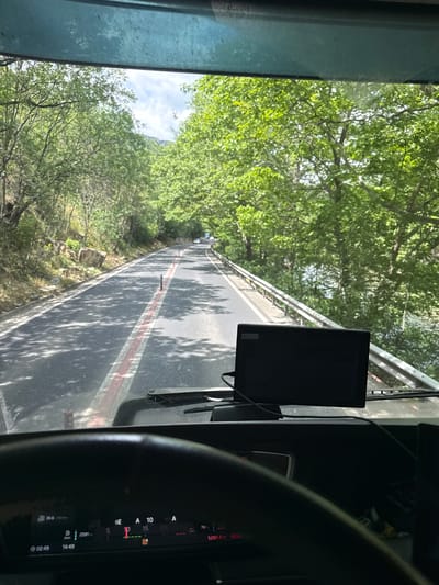 The image captures a view from inside a vehicle, likely a bus or truck, looking forward through the windshield onto a road winding through a verdant landscape. The vehicle appears to be moving along an elevated section of road or a bridge, bordered by a metal guardrail on the left. Lush green trees densely cover the areas beside and below the road, casting dappled shadows onto the asphalt. The sky, visible in patches through the foliage, is bright with some clouds, indicating a sunny day.
The road features distinct markings: a continuous red line running parallel to a broken white line, creating a unique lane division. In the foreground on the right, parts of the vehicle's interior are visible, including a portion of a steering wheel, a dark dashboard or instrument panel, and a small black monitor mounted to the windshield frame.
Notable details from the vehicle's instrument cluster include:
- "36.6" (possibly temperature or speed)
- "B 2591" (likely distance or mileage)
- A digital clock displaying "02:49" (possibly elapsed time or a secondary clock) and "14:49" (confirming the time of day as 2:49 PM).
- Indicators such as "A 10", "P 15", "A", and "70".
- A numerical reading "5281.4" and "500".
The scene suggests travel through a natural, possibly mountainous or hilly region, consistent with the location context of Stara Kresna, Bulgaria. The clear sky and strong shadows indicate bright, daytime conditions.
