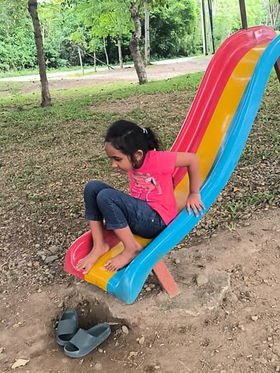 A young girl is positioned on a colorful playground slide in an outdoor setting in Lambarotunong, Indonesia. She is sitting mid-way down the slide, facing towards the right side of the frame with her head tilted slightly forward, looking down. Her expression appears focused or contemplative. She is barefoot, wearing dark blue jeans and a bright pink short-sleeved t-shirt with an indistinct graphic design. Her dark hair is pulled back into a ponytail.
The slide is made of plastic sections in red, yellow, and blue, appearing to be a standard children's playground slide. Its base is partially supported by a red block and some concrete. At the bottom of the slide, two dark grey or teal rubber slide-style slippers are neatly placed on the ground.
The ground around the slide is a mix of bare soil, small pebbles, and scattered patches of dry leaves and sparse green grass. In the background, a lush and dense area of green trees and foliage is visible, suggesting a natural or park-like environment. A dirt path or road can be seen winding through the trees in the distance. The lighting is soft and even, suggesting either an overcast day or early morning/late afternoon, as there are no harsh shadows. No discernible text is visible on any objects or in the background.