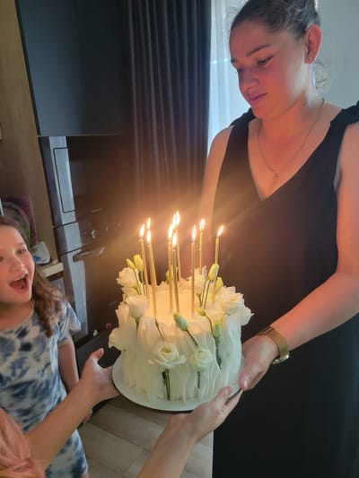 In Biaroza, Belarus, a woman in a black dress holds a white cake adorned with flowers and lit candles. To her left, a young girl with long brown hair in a blue tie-dye shirt looks at the cake with an open mouth. Another person's hands are visible reaching for the cake from the bottom left. The background shows dark curtains and a kitchen appliance.