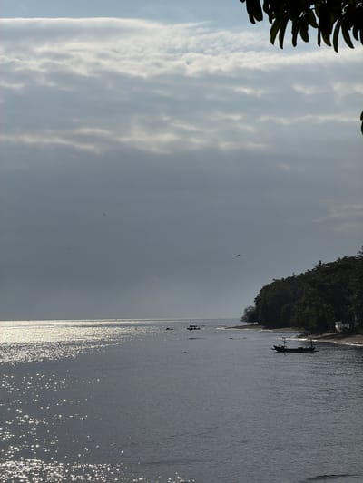 The image depicts a coastal scene at Pasir Putih, Indonesia, under a cloudy sky. The water is calm and reflects the light, creating a shimmering effect on the surface. Several small, dark-colored boats are visible on the water, some near the shore and others further out. The coastline is lined with lush green trees, with a sandy beach in between the water and the vegetation. The overall atmosphere is serene and natural.