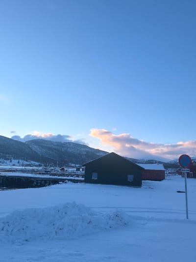 The image displays a winter landscape in the city of Gibostad, Norway. The foreground consists of a vast expanse of snow-covered ground, featuring a large, irregular pile of disturbed snow on the left. To the right, a circular traffic sign, indicating 'No Stopping' (blue disk with a red border and diagonal line), is mounted on a grey pole, emerging from the snow.

In the midground, a dark green, single-story building with a gable roof and two small rectangular windows on its visible side stands prominently. Behind it, slightly to the right, a smaller red building with a similar gable roof is visible. A body of water, likely a fjord, with visible pier structures and several moored boats, separates these buildings from the distant townscape.

The background features snow-covered mountains rising steeply. The mountain peaks are partially obscured by clouds, some of which exhibit a warm, golden-pink illumination, contrasting with the otherwise clear blue sky. No individuals are visible, and no discernible actions or interactions are occurring. The scene is static, illuminated by ambient daylight, possibly early morning or late afternoon.