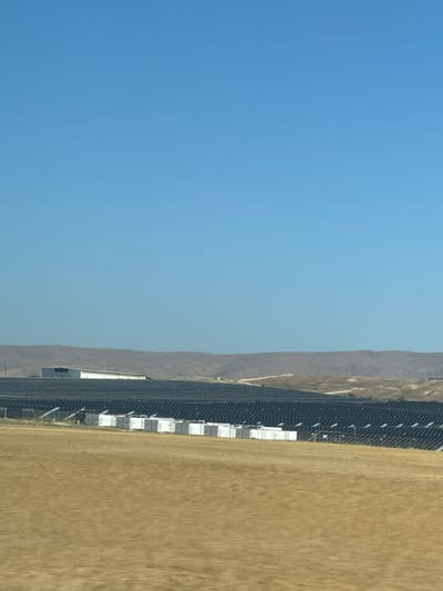 The image shows a landscape in Arad, Israel, under a clear blue sky. To the right is a field of dry, golden-brown grass. A long, dark fence runs between the field and the adjacent land. White rectangular structures are visible intermittently along the fence line. In the background, a low, arid mountain range can be seen.