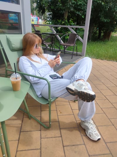 A young woman with long blonde hair and sunglasses is sitting in a green lounge chair on a patio. She is wearing a white long-sleeved shirt and light gray sweatpants, with white sneakers on her feet. She is looking down at her phone, which she is holding in her hands. A plastic cup with a brown beverage and a black straw sits on a small green table next to her.

The patio is paved with light brown square tiles. Behind the woman, there is a wooden deck with tables and chairs. Beyond the deck, there is lush green grass and trees. In the background, there appears to be a colorful inflatable structure, possibly a bouncy castle.

The scene suggests a relaxed daytime setting, likely outdoors at a cafe or a recreational area. The lighting indicates it's daytime, possibly late morning or afternoon, with good natural light. The woman's posture and focus on her phone suggest she is engaged in a casual activity, possibly browsing or communicating. The overall mood is one of leisure and relaxation. There is no visible text in the image.