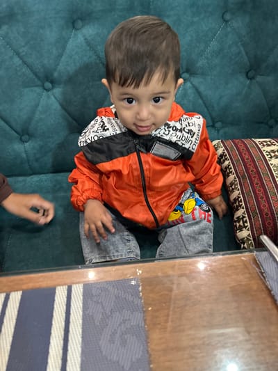 A child wearing an orange and black jacket and gray jeans sits on a teal, tufted couch. A decorative pillow rests behind the child. A hand of an adult is visible in the foreground. The location is inferred to be in or near Ақсу, Kazakhstan, based on the request.