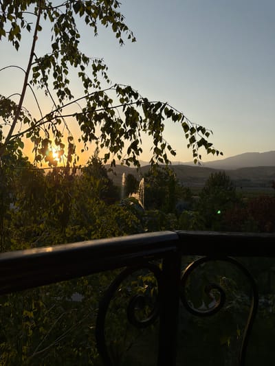 A serene sunset scene unfolds over a landscape in Karankultugay, Uzbekistan, viewed from a balcony. The foreground is dominated by the dark silhouette of a balcony railing with decorative scrollwork. Beyond the railing, lush green trees fill the midground, their leaves catching the warm, golden light of the setting sun. The sun itself is partially obscured by the branches and leaves of a tree in the upper left portion of the frame, casting a soft glow across the scene. In the distance, rolling hills fade into a hazy horizon under a pale blue sky. A fountain can be seen in the middle ground, its water catching the light. The overall atmosphere is peaceful and tranquil.