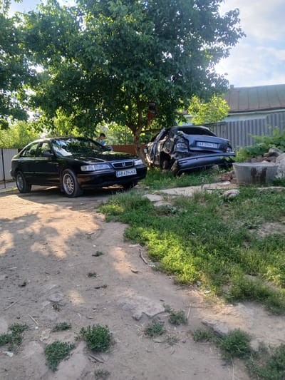 In the city of Pishchanka, Ukraine, an outdoor scene depicts two vehicles and a person. On the left, a black sedan, identified by the license plate AB 6394 KA, is parked on a dirt and gravel surface. To its right, a heavily damaged dark gray sedan, with license plate AA 0964 TC, rests at an angle, its rear severely deformed. The damaged car appears to be in a state of disrepair, possibly having been involved in an accident or experiencing a fire, evidenced by the broken rear window and mangled bodywork. A young person, partially obscured by the black car and the foliage of a large tree, stands near the vehicle on the left, appearing to be holding a blue object. The environment is characterized by lush green trees with dappled sunlight filtering through their leaves, a corrugated metal fence in the background, and patches of overgrown grass and vegetation. There are also some discarded bricks and a large concrete trough visible near the damaged vehicle. The scene suggests a residential or rural setting, possibly a yard or an informal parking area, given the presence of the fence and the unpaved ground.