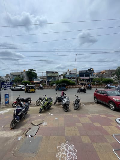 The image shows a street scene in Kurnool, India. The foreground features a tiled sidewalk with several parked motorcycles and scooters. A section of the sidewalk has a damaged manhole cover with debris around it. In the background, a busy street is visible with traffic including cars, auto-rickshaws, and motorcycles. Several commercial buildings line the street, with signage indicating businesses such as "DENNY'S DRESSES" and "FRIENDS A/C RESTAURANT". The sky is overcast with clouds, and numerous power lines crisscross the frame.