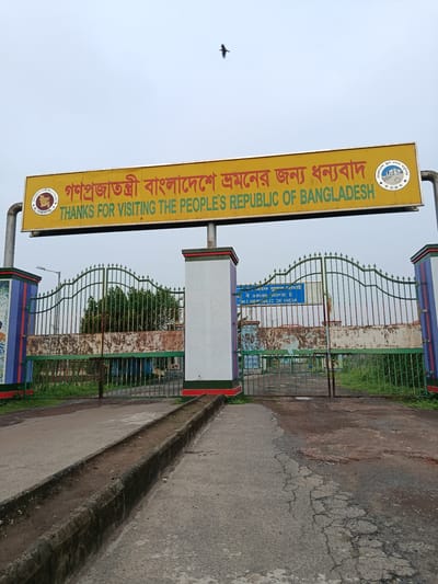 An overcast, daytime, eye-level exterior shot captures the Benapole border crossing in Bangladesh. The main subject is a large, yellow overhead sign with red text that reads "গণপ্রজাতন্ত্রী বাংলাদেশে শ্রমনের জন্য ধন্যবাদ" and "THANKS FOR VISITING THE PEOPLE'S REPUBLIC OF BANGLADESH". To the left of the text is the Bangladesh national emblem, and to the right is a circular logo with text. Below the sign is a green, ornate metal gate, partially rusted, with a white and colorful striped pillar on the left and a white pillar on the right. Beyond the gate, a building with a blue sign is visible, and greenery and trees surround the area. The road in the foreground is asphalt, cracked and worn, with a concrete drainage ditch running alongside it.