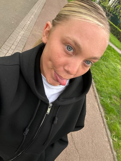 A light-skinned woman with blonde hair pulled back is sticking her tongue out and looking towards the camera. She has freckles and blue eyes. She is wearing a black hooded sweatshirt over a white shirt. The background consists of a paved walkway, a curb, and a grassy area. Based on available information and visual cues, the location is consistent with Noisiel, France.