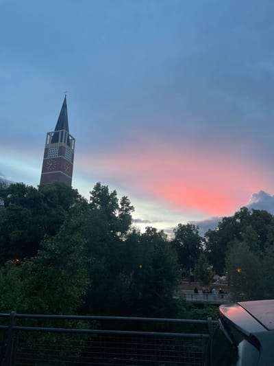 The image, taken in Pforzheim, Germany, presents a twilight scene dominated by a dramatic sky and a church tower. The upper sky transitions from a deep blue to a vibrant, soft pink and red cloud formation that extends horizontally, suggesting either sunrise or sunset. Below these colorful clouds, the sky fades into lighter gray and white hues.

A tall church tower with a dark, spired roof and a small cross at its apex stands prominently on the left. The tower itself is reddish-brown, accented with white or light-colored rectangular architectural details and features two visible clock faces with yellow markings; however, the precise time is not clearly discernible.

Dense, dark green trees fill the right side and lower portions of the frame, partially obscuring the tower. In the distant background on the right, through the foliage, some buildings and a few indistinct figures are visible, appearing to be seated or standing in an outdoor area. The foreground on the upper right includes a section of a car, possibly its roof or side mirror, reflecting the sky's colors, and a dark metal fence or railing with a grid pattern, indicating the photo was taken from an elevated vantage point. No visible text is legible within the scene.