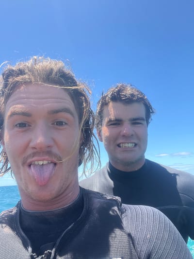Two men, their faces wet, pose closely against a bright blue sky and water backdrop. The man in front sticks out his tongue, while the other smiles widely, showcasing his teeth. Both are dressed in what appears to be wetsuits or similar dark attire. The angle of the shot gives a sense of being high up, possibly on a boat or cliff. Sunlight glistens on their wet hair and faces.