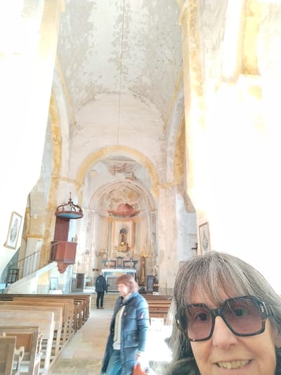 An interior shot of a church or cathedral, seemingly taken as a selfie. A woman with grey hair and large glasses smiles into the camera. Behind her, two other people are seen walking towards the altar. The interior features high ceilings with faded paintings, arches, a wooden pulpit, and rows of pews. The walls are aged, with peeling paint, suggesting a building of historical significance.
