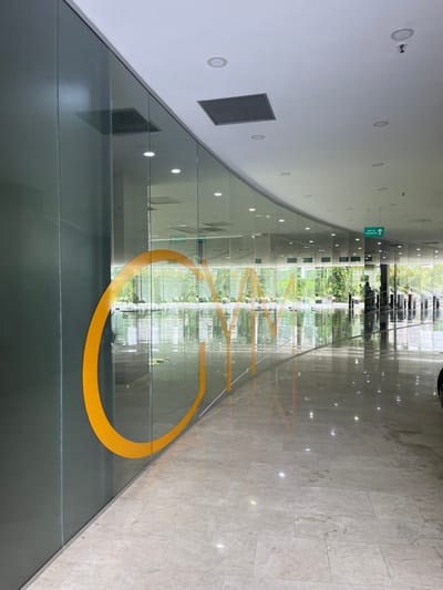 The image presents a ground-level view within the interior of the Gimnasio GYM Smart Fit Cañaveral, located in Floridablanca, Colombia. The dominant feature is a long, curved wall constructed primarily of clear glass panels, extending from the left foreground towards the right background. Prominently affixed to the lower half of this glass wall are large, stylized letters in a bold orange color, spelling "GYM". To the immediate left of the "GYM" lettering, a segment of the wall is opaque and dark grey. The floor is paved with large, light-toned, highly polished marble or ceramic tiles, exhibiting significant reflections of the overhead lighting and the visible outdoor environment. The ceiling is white, fitted with numerous recessed circular light fixtures and at least two square ventilation panels. Further along the corridor to the right, a green emergency exit sign, bearing the text "SALIDA DE EMERGENCIA" and a right-pointing arrow, is suspended from the ceiling. Through the transparent glass wall, an outdoor area is visible, characterized by dense green foliage, trees, and what appears to be a paved or concrete surface with some indistinct structures. A partial view of a dark, indistinct object is visible on the far right edge of the frame. No individuals are present, and no active interactions or actions are depicted within the scene.