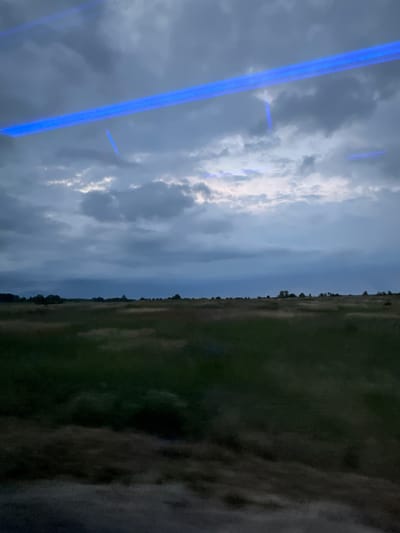 The image depicts a dimly lit, expansive field under a cloudy sky. The sky is characterized by dark, heavy clouds with streaks of lighter, diffused light breaking through in places. A distinct horizontal blue light bar, likely from an artificial source, cuts across the upper portion of the frame, casting reflections. Below the sky, a vast expanse of green and brown vegetation covers the landscape, appearing somewhat blurred, suggesting movement or a low-resolution capture. In the distance, a faint treeline marks the horizon, with scattered darker shapes that could be trees or shrubs. The overall impression is one of a natural landscape observed from a moving vehicle, possibly at dusk or under overcast conditions, with the addition of an artificial light element. The location in Chociule, Poland, is not visually discernible from the image itself.