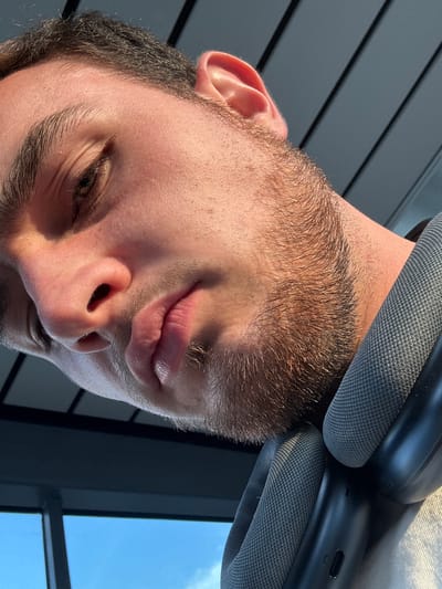 A close-up, low-angle shot features a young man's face, angled downwards, with his eyes partially closed. He has light brown hair, light skin, and a light stubble beard. The man is wearing dark gray headphones around his neck. The background consists of dark horizontal lines, suggestive of blinds or slats, and a bright blue sky with scattered clouds visible through a window. The lighting suggests it is daytime, with sunlight casting shadows on the man's face. There are no identifiable landmarks or features that would indicate a specific location within Apodaca, Mexico.