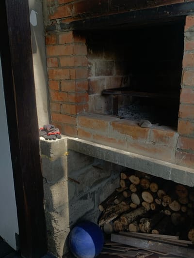 The image depicts an outdoor brick-built oven or fireplace structure located in Yasinia, Ukraine. The structure is primarily constructed from red bricks with visible mortar lines and features concrete elements for hearths and support shelves. The main firebox, centrally positioned, is dark inside with visible ash residue on its base and a dark metal grate or shelf spanning its width. A small, light-colored stone rests on the brick hearth within the firebox.

To the lower left, a pair of gray and red work gloves are placed on a concrete ledge. Below this ledge, the structure forms an open storage area where a stack of cut logs and smaller wood pieces are neatly arranged. A blue spherical ball rests on the ground at the base of the concrete support column on the far left. Adjacent to the brick structure on the left, a dark wooden beam or post is visible, bordering a light-colored, possibly plastered, wall section. Sunlight casts shadows, indicating an outdoor setting during daytime. No people are present, and no active processes are occurring.