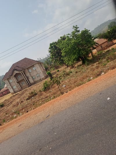 The image depicts an outdoor scene in Ochadam, Nigeria, captured from a slightly elevated perspective looking across a paved road towards structures and a hilly landscape. The lower right portion of the frame features a dark grey asphalt road, bordered on its left by a reddish-brown dirt shoulder where a small, dark, elongated object (possibly debris or a stick) lies.
Across the road, occupying the left midground, stands a multi-story, light-colored (possibly beige or cream) building with a prominent brown tiled roof. This building features multiple windows, many with visible grilles, and two distinct arched porticos or balconies on its front facade, indicating multiple levels. The ground surrounding this structure is largely undeveloped, covered in dry, light brown vegetation and sparse, smaller green plants.
Further into the midground, to the right of the primary building, scattered green trees and shrubs are visible. Behind these trees, a smaller, single-story building with a reddish-brown roof can be partially discerned. The background consists of rolling hills covered in light green foliage under a pale blue sky with diffused white clouds. Several dark utility lines traverse diagonally across the upper portion of the sky and tree line. No individuals, overt actions, or interactions are present within the scene. The overall environment suggests a semi-developed area with natural terrain features.