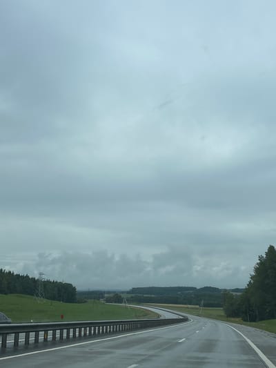 This image depicts a wet, curved highway in a rural, possibly forested area. The sky is overcast with heavy clouds. A guardrail runs along the left side of the road, with a white line separating the lane. In the distance, there are rolling green hills and a dense line of trees. A transmission tower is visible on a grassy slope to the left. The road surface appears reflective, indicating recent rain. The location is described as Lahojsk Selsaviet, Belarus. There are no people visible in the image.