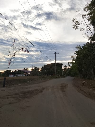 This is a photograph of a rural road in Nangrhoe Timu, Indonesia. The road is unpaved and appears to be dirt and gravel. Overhead, power lines stretch across the frame, supported by poles. To the left of the road, there is a field with some dry vegetation and a few scattered trees. In the distance, a building with a red roof can be seen, along with more trees and a transmission tower. To the right of the road, a dense line of trees stands tall. The sky is cloudy and overcast, suggesting it might be late afternoon or early morning. There are no people visible in the immediate foreground, but there might be a few individuals in the distance near the building on the left. Some small, red fabric pieces are tied to a thin tree branch on the left side of the image.