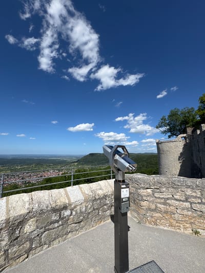 The image depicts a view from the Neuffen Castle ruins in Neuffen, Germany, on a bright, sunny day. In the foreground, a coin-operated viewer stands on a paved surface next to a stone wall. The viewer is a silver, binocular-like device mounted on a dark gray pedestal. The pedestal has various labels and a coin slot. The stone wall, constructed from rough-hewn, light-colored stones, forms a parapet overlooking a town and a valley. Beyond the wall, a metal railing runs horizontally, separating the viewpoint from the town below. The town of Neuffen is visible in the middle ground, characterized by red-tiled roofs and dense greenery. The landscape extends into the distance, with rolling hills covered in forests and fields under a vibrant blue sky dotted with white, fluffy clouds. To the right of the image, a section of the castle ruins, a circular stone tower, and more stone walls are visible, with trees growing above them. The overall scene conveys a sense of historical significance and scenic beauty.
