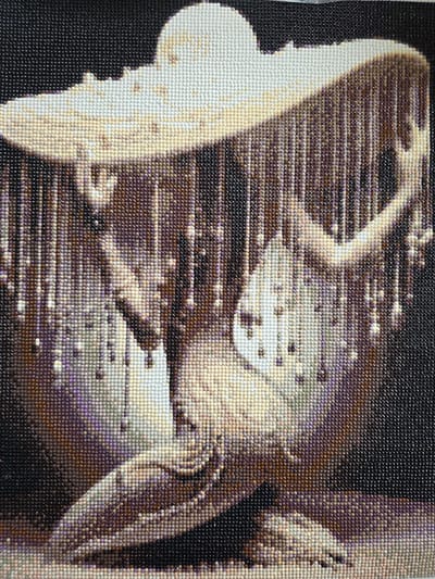 The image is a close-up of a diamond painting. The painting depicts a large, ornate hat with a brim casting shadows. From the brim, numerous strands of beads or diamonds hang down, creating a curtain-like effect. Beneath the hat, a figure is partially visible, appearing to be kneeling or in a seated position. The figure's form is indistinct, rendered in shades of beige, brown, and purple, blending with the shadows and the beaded strands. The background is solid black, providing a stark contrast that emphasizes the detailed textures of the hat and the figure. There are no discernible elements that would place this image in Claremorris, Ireland.