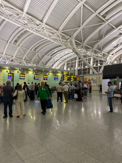 An indoor airport scene in Dar El Beïda, Algeria, shows a queue of travelers at a check-in counter. The ceiling features a complex white metal truss system supporting corrugated metal panels. The floor is a polished gray tile. People, dressed in casual attire, stand in line, some with luggage. Behind them, illuminated check-in counters display flight information on screens, including flight numbers and airline logos such as Lufthansa. Signs in Arabic and English are visible on the wall. One man in the foreground wears a green jacket and holds a light blue cooler and a dark blue bag. Another man in a light blue polo shirt and dark pants stands to the right, facing away from the camera. The overall lighting is bright and artificial.