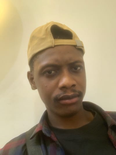 A man is featured in a close-up portrait, oriented vertically. He has dark skin and short facial hair, including a mustache and chin beard. He looks directly into the camera with a neutral expression. He is wearing a light tan or beige baseball cap backward, a dark t-shirt, and a plaid shirt with patterns of red, blue, and black over it. A dark strap, possibly from a bag, is visible over his right shoulder. The background consists of a plain, light-colored wall, suggesting an indoor setting. The lighting is soft and ambient. No visible text is present in the image. The location context is Animashawun, Nigeria.