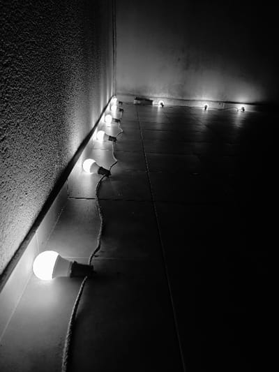 A black and white photograph depicts an interior corner located in Villa San Andrés, Argentina. Approximately twelve illuminated LED-style light bulbs, each with a white frosted dome and a dark base, are positioned linearly along the base of a wall on a dark, square-tiled floor. The bulbs are interconnected by a dark, flexible electrical cord that snakes between them. The wall on the left features a rough, textured surface, while the wall beyond the corner appears smoother but also worn. The scene is dimly lit, with the light bulbs serving as the sole visible light source, casting localized illumination and deep shadows that extend towards the right and into the distant corner. No individuals or other subjects are present.