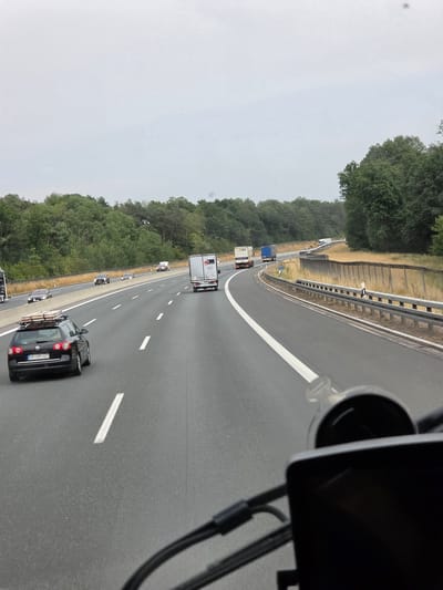 The image shows a view from inside a vehicle on a highway, likely the A3 Autobahn, near Bischbrunn, Germany. The weather is overcast. In the foreground, part of the vehicle's interior, including a dashboard and possibly a camera mount, is visible. The highway has multiple lanes, indicated by white lines. Several vehicles are on the road: a black station wagon with a load of wood on its roof is in the left lane, followed by a white car and a white truck. In the right lane, further ahead, are two trucks, one white and one blue, and a few other cars. The highway is bordered by a concrete barrier and a dry, grassy embankment with a wooden fence. Beyond the embankment, a dense forest of green trees stretches to the horizon under the cloudy sky. The scene depicts typical highway traffic in a rural, wooded area.