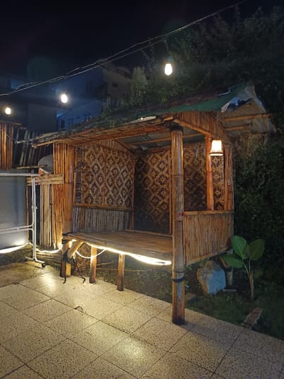 The image depicts a nighttime outdoor scene in Palasari, Indonesia, focusing on a raised bamboo structure. The structure, approximately 2.5 meters wide and 2 meters deep, is a traditional-style gazebo or hut supported by bamboo poles. Its three visible walls are constructed from woven bamboo mats featuring a repeating diamond pattern. The roof is composed of natural materials, possibly thatch, overlaid with a light blue or green tarpaulin. A horizontal string of at least four exposed bulb lights hangs overhead, partially illuminating the scene.

A single lantern with a woven shade is affixed to an interior bamboo post of the structure. A strip of white LED lights is draped along the underside perimeter of the raised bamboo plank floor. The ground in the foreground consists of light-colored, textured tiles. To the immediate left of the structure, a separate bamboo fence and a white metal utility frame on wheels are present. To the right of the structure, a small patch of grass contains a broad-leafed plant and two irregular rocks. In the distant background, dimly lit multi-story buildings are visible against the dark night sky. No individuals, animals, or specific activities are depicted.