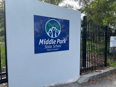 The image displays an outdoor scene, likely at the entrance or boundary of a school in Riverhills, Australia, though it is currently rotated 90 degrees clockwise. On the right, a light grey concrete wall or pillar serves as a backdrop for a prominent rectangular blue sign. This sign clearly identifies the institution as "Middle Park State School" with white lettering, and includes the motto "'Each to Succeed'". Below the school's name, a circular logo features three stylized white figures within a green leafy wreath, set against the blue background.

To the left of the grey wall, a black metal fence extends horizontally across the frame. Behind the fence, lush green trees and foliage are visible, with some scattered brown leaves and debris on the ground near a paved or curbed area at the top of the rotated image. Attached to the fence on the far left is a white poster, which, despite being partially obscured and blurry, displays several smiling faces. Identifiable text on the poster includes "Supporting our students" and a logo resembling "WM".

The scene appears to be well-lit, indicating it is daytime. No people or specific activities are taking place; it presents a static view of the school's perimeter.