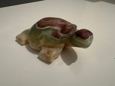 A carved stone turtle figurine, with swirls of green, white, and red, sits on a white surface. The lighting casts a shadow to the right of the figurine. The background is dark grey. The image was taken in Stoke-on-Trent, United Kingdom.