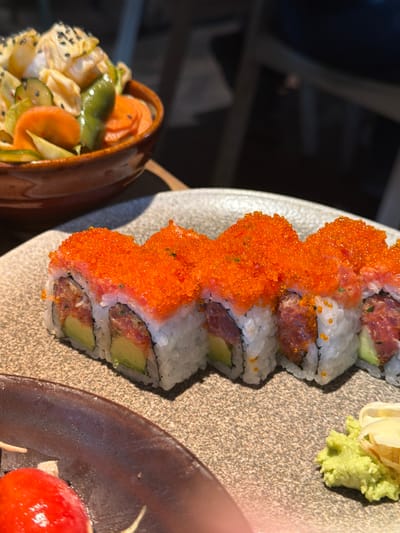 The image depicts a close-up view of food arranged on a table within a restaurant setting in Kiryat Ono, Israel. The primary subject is a line of six sushi rolls on a light brown, textured plate. Each sushi roll is an uramaki type, featuring white rice on the exterior, filled with dark red chopped fish (likely tuna) and sliced avocado, and generously topped with small, bright orange tobiko or masago roe. To the bottom right of the sushi plate, a small portion of green wasabi paste and several slices of pickled ginger are visible. In the mid-ground, to the left, a small, dark brown bowl contains a salad or pickled vegetables, including sliced carrots, green peppers or cucumbers, and light-colored vegetables topped with black sesame seeds. A partial view of another dark brown plate is visible in the bottom left corner, holding a small red, spherical item. The background is blurred, showing indistinct dark shapes consistent with restaurant furniture like chairs and tables. No individuals are visible.