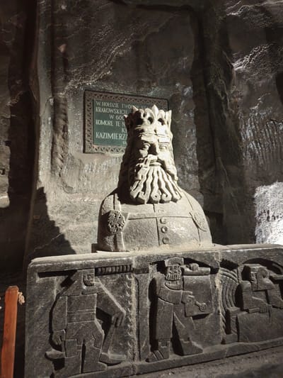 The image captures a stone statue bust of a crowned figure, likely a king, located in the Wieliczka Salt Mine in Wieliczka, Poland. The bust is placed atop a rectangular stone block adorned with carved figures. A plaque hangs on the stone wall behind the statue. The environment is an underground chamber with textured stone walls. The lighting is dim, creating highlights and shadows.