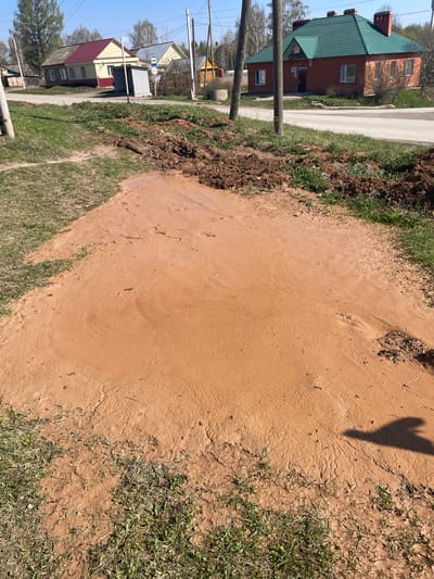 The image depicts a flooded area in Поедуги, Russia. The ground is covered in a thick layer of reddish-brown mud, obscuring the grass and extending towards the roadside. A pile of excavated soil sits on the edge of the water. In the background, there are houses and a bus stop. An electric pole is close to the houses. The shadow of a person/object can be seen over the mud.