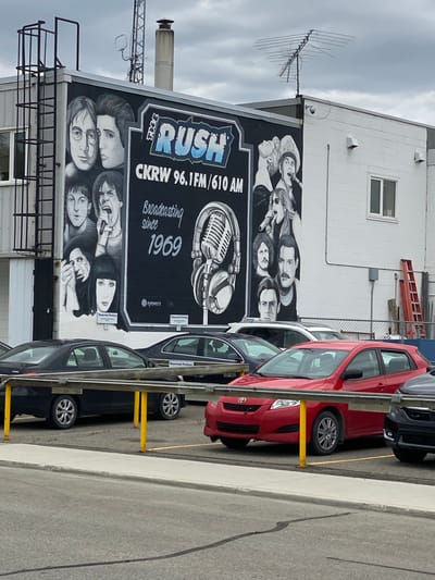 The image depicts a mural on the side of a building in Whitehorse, Canada, which features portraits of musicians and a vintage microphone with headphones. The mural is for "The Rush CKRW 96.1FM / 610 AM," indicating it's likely the exterior of a radio station. The text below reads "Broadcasting since 1969." Several parked cars, including a red Toyota and a black sedan, are visible in the foreground, with a yellow barrier separating the parking lot from a walkway. The building appears to be a commercial structure with a ladder attached to its side. The sky is overcast.