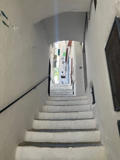 A white stone staircase ascends through a narrow alleyway in Pogerola, Italy, flanked by white stucco walls. The staircase has dark edges and is partially shaded by an overhead archway. A black metal handrail runs along the left side of the stairs. On the left wall, two dark green fish silhouettes are affixed. The alley opens up to a sunlit area at the top, revealing glimpses of buildings with green shutters and a distant lantern. A black metal grate is visible on the right wall. The scene is devoid of any people.