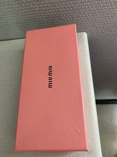 A close-up, top-down shot shows a salmon-pink, rectangular box with the embossed black lettering "MIU MIU" vertically oriented on its surface. The box has a textured, leather-like finish with visible creases and a small scratch near the bottom left corner. It rests on a light gray, textured surface, possibly a countertop or table. Behind the box, a dark gray, woven material is visible, suggesting a wall or upholstery. The lighting is diffused, casting no strong shadows. There are no people or animals present in the image. The scene appears to be indoors, within a domestic or retail setting. The image does not provide any information about the location in the city of Maksimkovo, Russia.