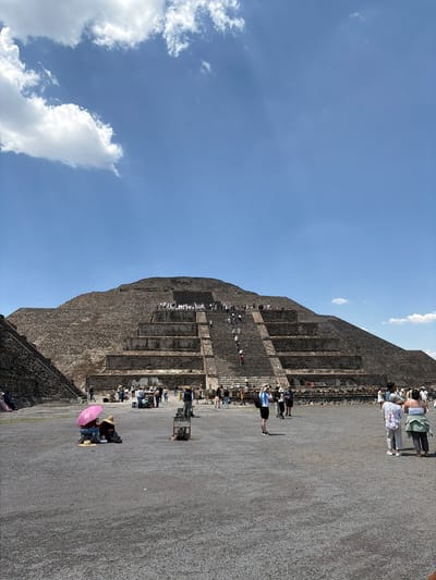 The image shows the Pyramid of the Sun in Teotihuacan, located in San Francisco Mazapa, Mexico. The pyramid, constructed of layered stone, is covered in gray-brown weathered material. Numerous people are seen climbing and standing on the pyramid's steps, and others are on the ground around the base. The ground near the pyramid appears to be gravel or compacted earth. The sky is blue with scattered white clouds.