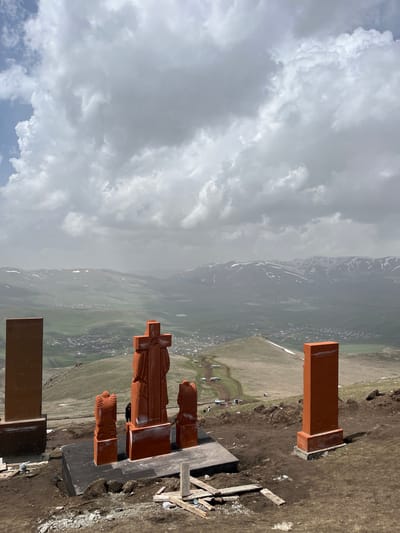 The image shows the Vardapet Memorial in Hartagyugh, Armenia. The Vardapet Memorial features a large, rust-colored cross flanked by stylized figures. The cross and figures are set upon a gray platform. Nearby are rectangular blocks, also rust-colored. The background shows a landscape with mountains and a sky filled with clouds. Buildings are visible in the valley below. The area around the memorial appears to be dirt and grass. A few pieces of wood debris are also visible.