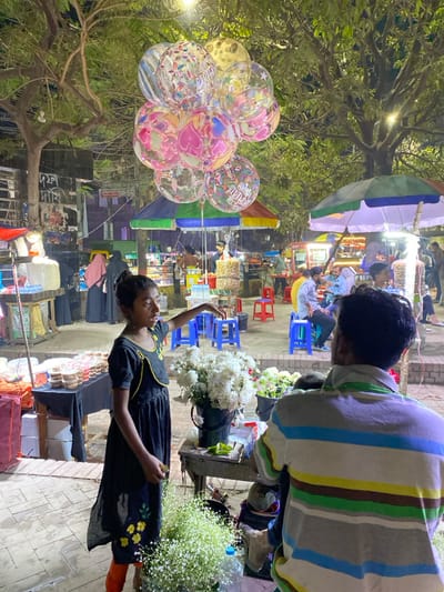 The image captures an illuminated outdoor night market scene in Narayanganj, Bangladesh. In the foreground-center, a young girl, dressed in a black, short-sleeved dress with yellow accents and orange leggings, stands facing left with her left arm extended and slightly raised, appearing to point or gesture. She holds a small yellow object in her right hand. To her immediate right, a black bucket filled with white flowers, possibly chrysanthemums, rests on a small wooden table. Below this, a clear plastic bottle contains a large quantity of white baby's breath flowers.

In the foreground-right, the back of a man wearing a horizontally striped polo shirt (grey, white, brown, blue, green, yellow) is visible, facing away from the viewer. A child's head is partially visible near his left shoulder.

Approximately 10-12 clear, spherical balloons with various colorful patterns, including hearts, stars, polka dots, and "Happy Birthday to you" text, float prominently in the upper center of the frame.

The midground features several street vendor stalls, each covered by a large, colorful striped umbrella (blue, green, white, red). These stalls display various food items, including what appear to be puffed rice snacks in transparent containers. Multiple individuals are visible around these stalls, some seated on blue and red plastic stools.

The background consists of large trees with visible foliage and branches, illuminated by overhead streetlights. On the far left, portions of buildings are visible, one featuring posters or signs with Bengali script. The ground is paved with light-colored tiles or bricks. The scene depicts a busy commercial environment after dark.