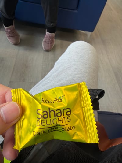 The image depicts an indoor scene with two individuals. In the foreground, a person is seated, wearing light grey sweatpants. Their left hand holds a crinkled, bright yellow foil-wrapped chocolate bar, branded "Abu Auf Sahara DELIGHTS Dark chocolate with Almonds." To the right of this person's leg, a dark, textured strap, possibly from a bag, and a small brown leather-like accent are visible. In the background, opposite the foreground individual, another person is seated, with only their lower legs and feet visible. They are wearing dark trousers or leggings, black socks, and light pink or mauve athletic shoes with laces. The flooring is composed of light grey, wood-effect planks. A portion of dark blue upholstered furniture, likely a sofa or chair, is visible between the two individuals in the background. The specific location within Connah's Quay, United Kingdom, cannot be determined from the visual information provided.