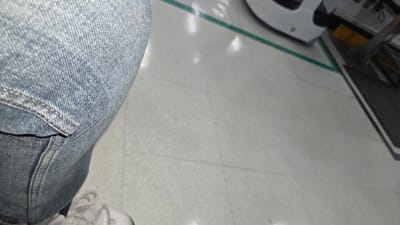 The scene depicts a partial view of an individual standing on a light grey tiled floor at a location in La Tijera, Mexico. The person's left leg is visible in the foreground, clad in light blue denim jeans with visible contrast stitching on a seam or pocket. A white athletic shoe with white laces is visible on the individual's foot at the bottom of the frame. The tiled floor features a distinct green line, approximately 2-3 inches wide, extending diagonally from the upper left towards the middle right. Beyond this green line, a white, curved object, possibly the front section of a vehicle or piece of equipment, is partially visible and out of focus. Further to the right, a rectangular dark grey or black floor mat is positioned on the floor, adjacent to what appears to be a metallic structure. Reflections from overhead light sources are evident on the glossy tiled surface. No direct interactions or specific actions beyond standing are discernible.
