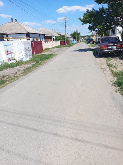 The image depicts a quiet, paved road in the village of Zaluzhnoye, Russia, on a sunny day with a blue sky and scattered clouds. The road is lined with single-story houses and fences. On the left side of the road, stacks of white building materials, possibly insulation or foam blocks, are visible with "Block" branding. Behind the materials, a red fence and the side of a house with a satellite dish are seen. Further down the road on the left, several houses with grey, corrugated roofs and various colored fences are visible. On the right side of the road, parked cars are present, including a dark-colored sedan and a reddish-brown sedan with the license plate "C 210CT 36". A large green tree shades a portion of the right side of the road and a white house. Green grass and weeds grow along the sides of the road and in front of the houses. A utility pole with wires is also visible in the distance on the left. The road surface is asphalt, with some white chalk-like markings visible in the foreground.