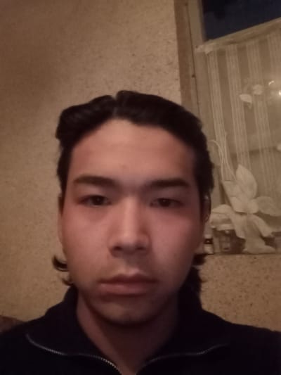 A male individual is depicted in a close-up, indoor photograph, identified as being in Takhtakuprik, Uzbekistan. The subject has dark, medium-length hair, centrally parted and pulled back. Apparent redness and minor swelling are visible under both eyes and on the upper cheekbones. He wears a dark, possibly navy blue, zippered jacket or shirt. The background features a light brown, textured wall on the left. To the right, a window is visible, covered by a sheer, light-colored curtain with a floral pattern. The exterior visible through the upper portion of the window appears dark, suggesting low light conditions. On a surface beneath the window, a small bottle with a red cap and other indistinct items are present. The scene is illuminated by dim, artificial light.