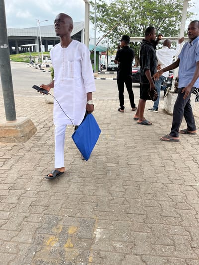 The image captures an outdoor scene in Gwaska, Nigeria, featuring several men on a paved walkway during what appears to be an overcast daytime.
In the foreground, a man wearing a white long-sleeved tunic with subtle embroidery and matching white trousers stands, holding a black smartphone in his left hand and a blue bag or folder in his right. He wears black slide sandals and a watch. His head is bald, and he is looking to his right.
Behind him and to the left, a group of other men are visible. One man in a light blue collared shirt and dark trousers stands near a pole, while another in a black short-sleeved shirt and black shorts holds a dark hat. Further back, another man in a white shirt and jeans is partially visible, and a man in a black shirt and black trousers, wearing a dark baseball cap, is also present. Most of the men are wearing sandals or slippers.
The setting is an urban or semi-urban environment with a patterned interlocking brick pavement. Green foliage from trees is visible on the left side of the frame, and modern buildings with large windows and gray/white facades are in the background. Partially visible cars are parked in the midground. The men appear to be standing around, possibly conversing or waiting. There is some unreadable text on a distant sign.