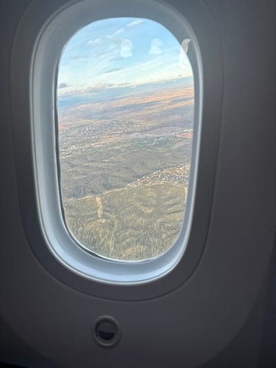 The image displays an aerial view from an aircraft window, showing a diverse landscape near Santo Domingo, Dominican Republic. The light grey, oval-shaped airplane window frame dominates the foreground, with a circular control knob, possibly for dimming, visible on the lower interior panel.
Through the window, the ground below features a mosaic of dense green vegetation and forested areas interspersed with numerous residential developments characterized by clusters of buildings. Further into the distance, the terrain transitions to lighter brown and yellowish hues, indicative of more open land, possibly agricultural fields or less vegetated areas, extending towards a hazy horizon. The sky above is light blue with scattered white clouds. No people or direct interactions are visible. The scene is illuminated by natural daylight.