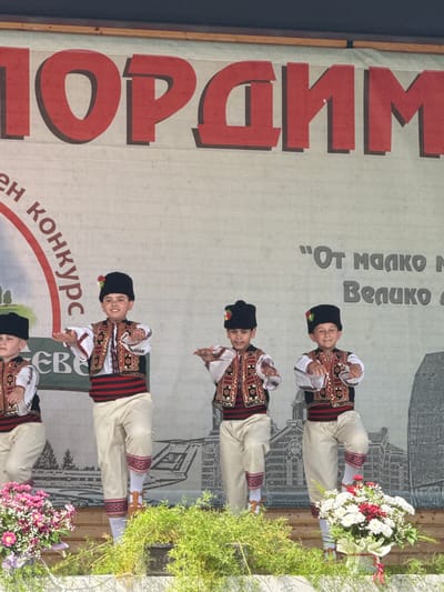 A group of four young boys in traditional Bulgarian folk costumes are performing on an outdoor stage. They are wearing black hats adorned with flowers, white shirts with embroidered vests, red and black striped sashes, and cream-colored pants with decorative red trim around the ankles. The boys are in mid-dance, with their arms outstretched and their legs positioned in a dynamic pose. Behind them is a large banner with red lettering and a faint image of buildings. The banner appears to read "PORADIM" in large red letters at the top. To the left of the boys, a circular emblem with text is visible on the banner. To the right, there are two lines of text in a grayish hue. The stage itself is made of wood, and in front of it are potted plants and bouquets of pink and white flowers. The overall scene suggests a cultural event or festival taking place in the city of Pordim, Bulgaria.