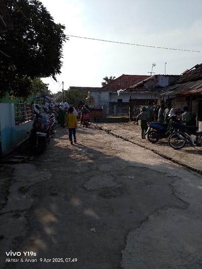 The image shows a narrow street in Kecamatan Gegesik, Indonesia, with a group of people, mostly children, walking and riding bicycles. There are motorcycles parked on both sides of the street, and the street is covered in uneven concrete. The houses on either side of the street are a mix of modern and traditional architecture. The weather is clear and sunny, and the sky is blue with some white clouds. Some shadows cast by the buildings, trees and people are on the ground. There are two buildings in the background, one with a partially collapsed roof and one with a white painted wall. The scene is one of ordinary life in a rural Indonesian village. The image was taken on April 9, 2025, at 7:49 AM.