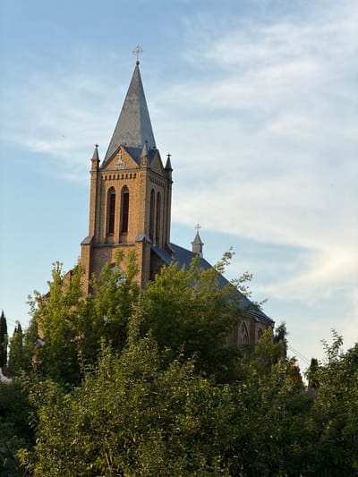 The image captures a brick church with a tall, pointed spire, partially obscured by lush green trees in the foreground. The church's facade is made of light brown bricks, with arched windows and decorative architectural elements. A cross is visible atop the spire and on a smaller turret. The sky is a pale blue with wispy white clouds. The overall impression is that of a serene, somewhat aged religious building set against a natural backdrop. The scene is likely located in Красненскі сельскі Савет, Belarus, given the context.